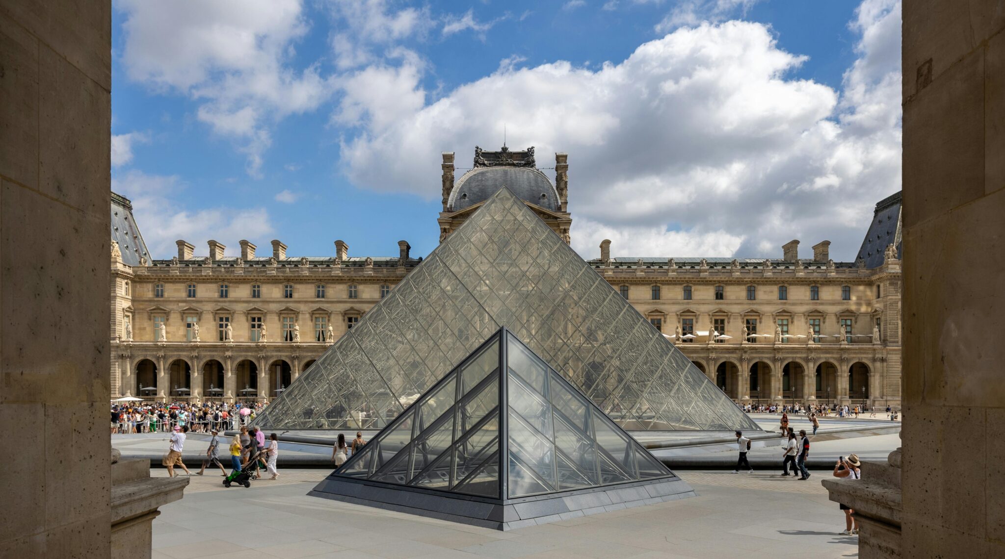 Visitors explore the Louvre Museum courtyard and admire the glass pyramid under a sunny sky